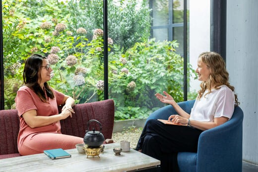 Klientin im lachsfarbenen Kleid und Coach in weißer Bluse sitzen sich gegenüber vor einem Fenster und sprechen darüber wie Klientin lernen kann, ihre negativen Gedanken auch im Alltag zu stoppen.